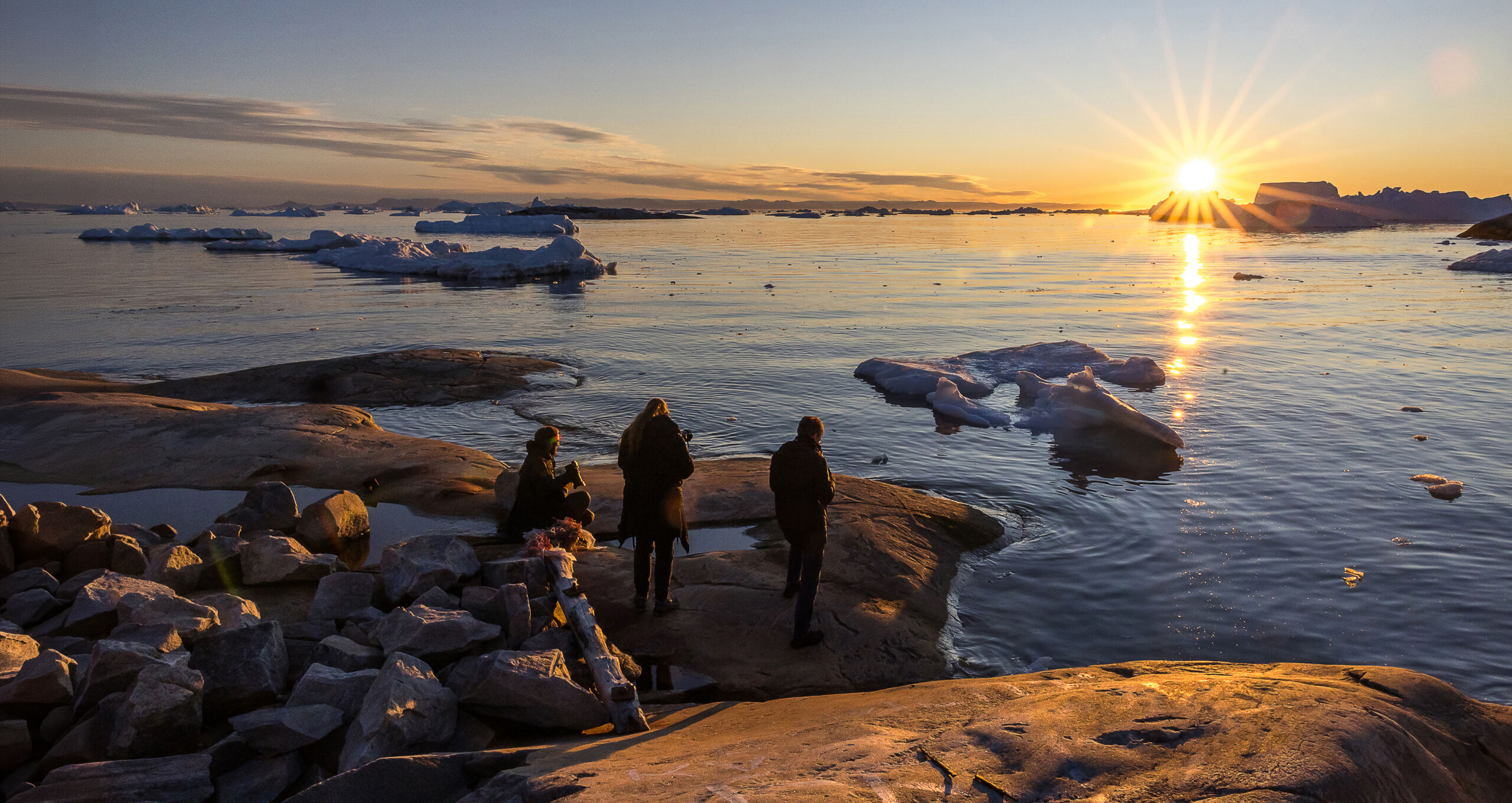 Drei Menschen fotografieren Eisberge im goldenen Abendlicht am felsigen Ufer in Grönland.