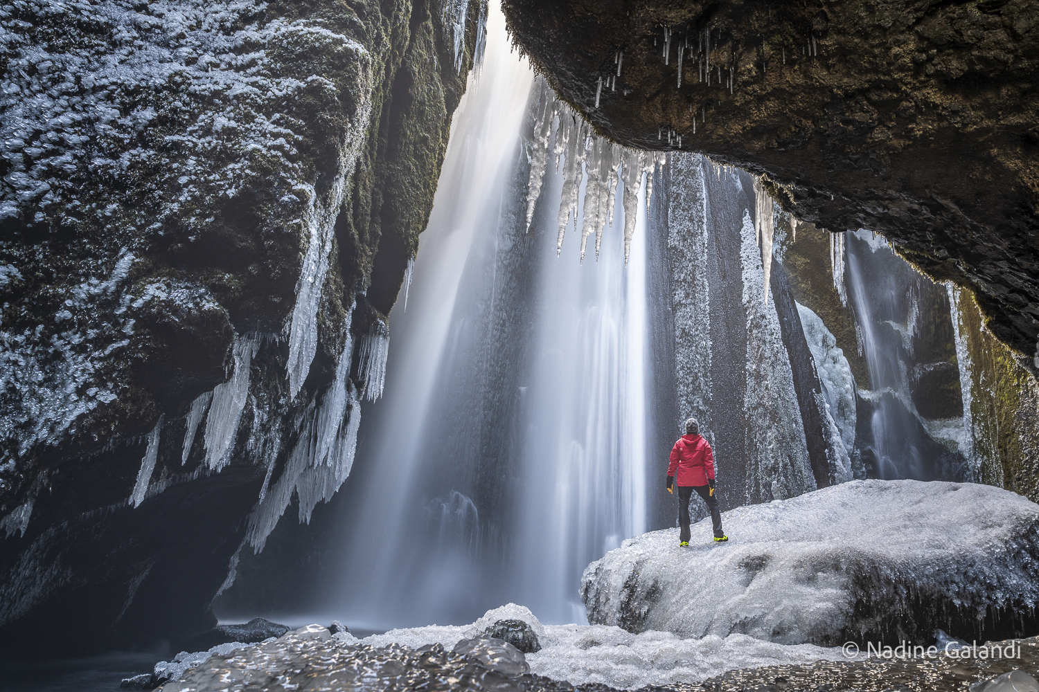 Gljúfrabúi Wasserfall im Winter – Blick hinter den Wasserfall Person im roten Anorak hinter dem Gljúfrabúi Wasserfall in Island, Eiszapfen und Wassernebel im Winter