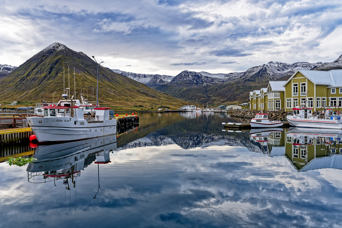 Boote im Hafen von Siglufjörður, Nordisland – spiegelglattes Wasser, bunte Holzhäuser und schneebedeckte Berge im Hintergrund.