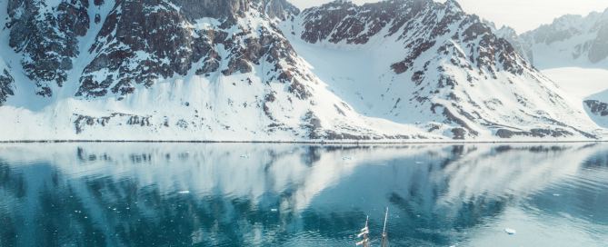 Segelschiff Ocean Sherpa im Scoresbysund in Ostgrönland – Spiegelung der schneebedeckten Berge im türkisblauen Wasser unter klarem arktischem Licht.