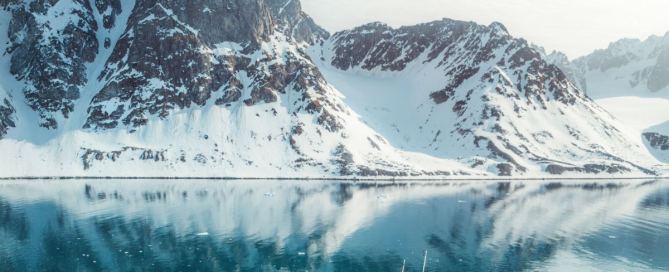Segelschiff Ocean Sherpa im Scoresbysund in Ostgrönland – Spiegelung der schneebedeckten Berge im türkisblauen Wasser unter klarem arktischem Licht.