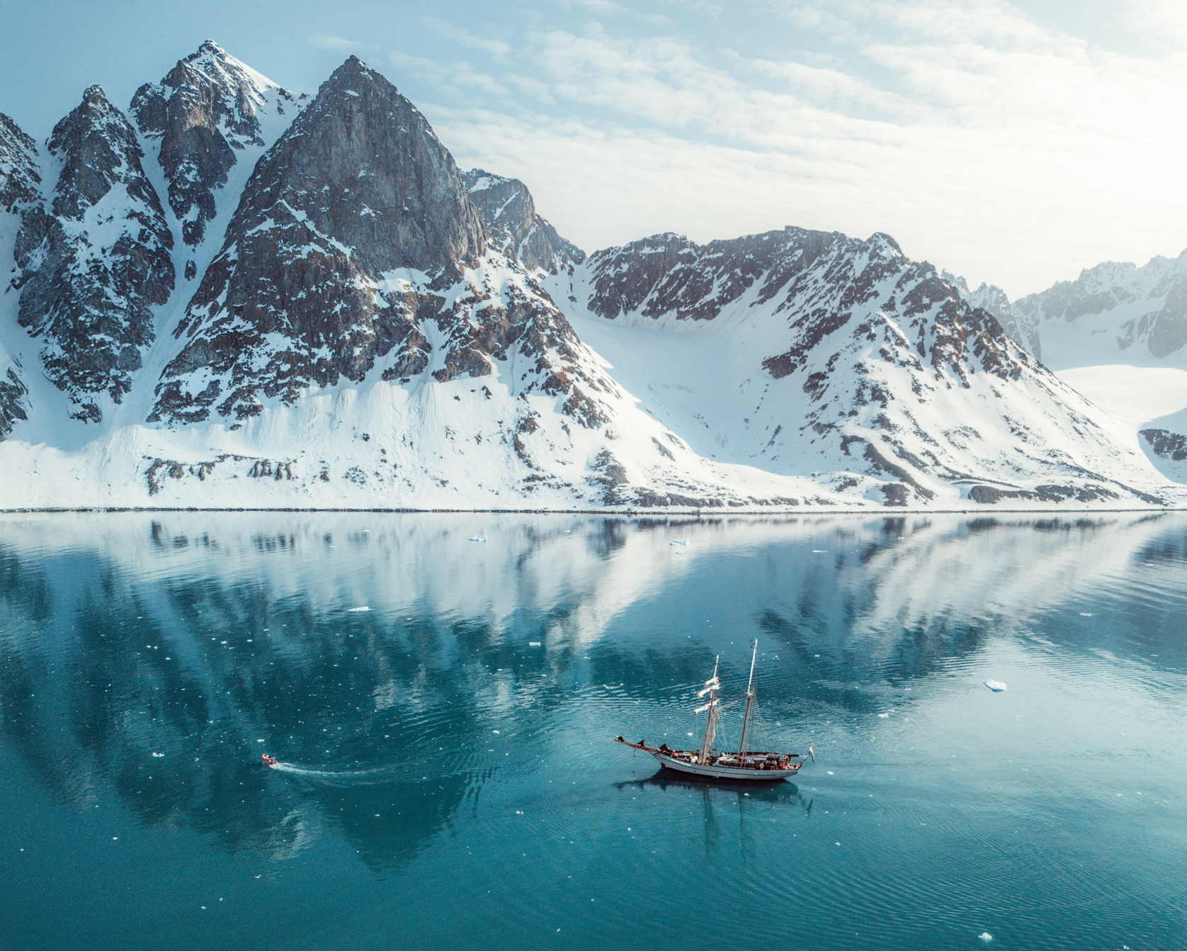Segelschiff Ocean Sherpa im Scoresbysund in Ostgrönland – Spiegelung der schneebedeckten Berge im türkisblauen Wasser unter klarem arktischem Licht.