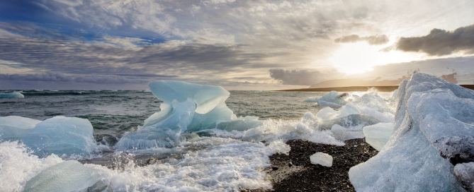 Eisbrocken am Diamond Beach in Island, Wellen brechen über schwarzen Kies, tief stehende Sonne und dramatischer Himmel im Hintergrund.