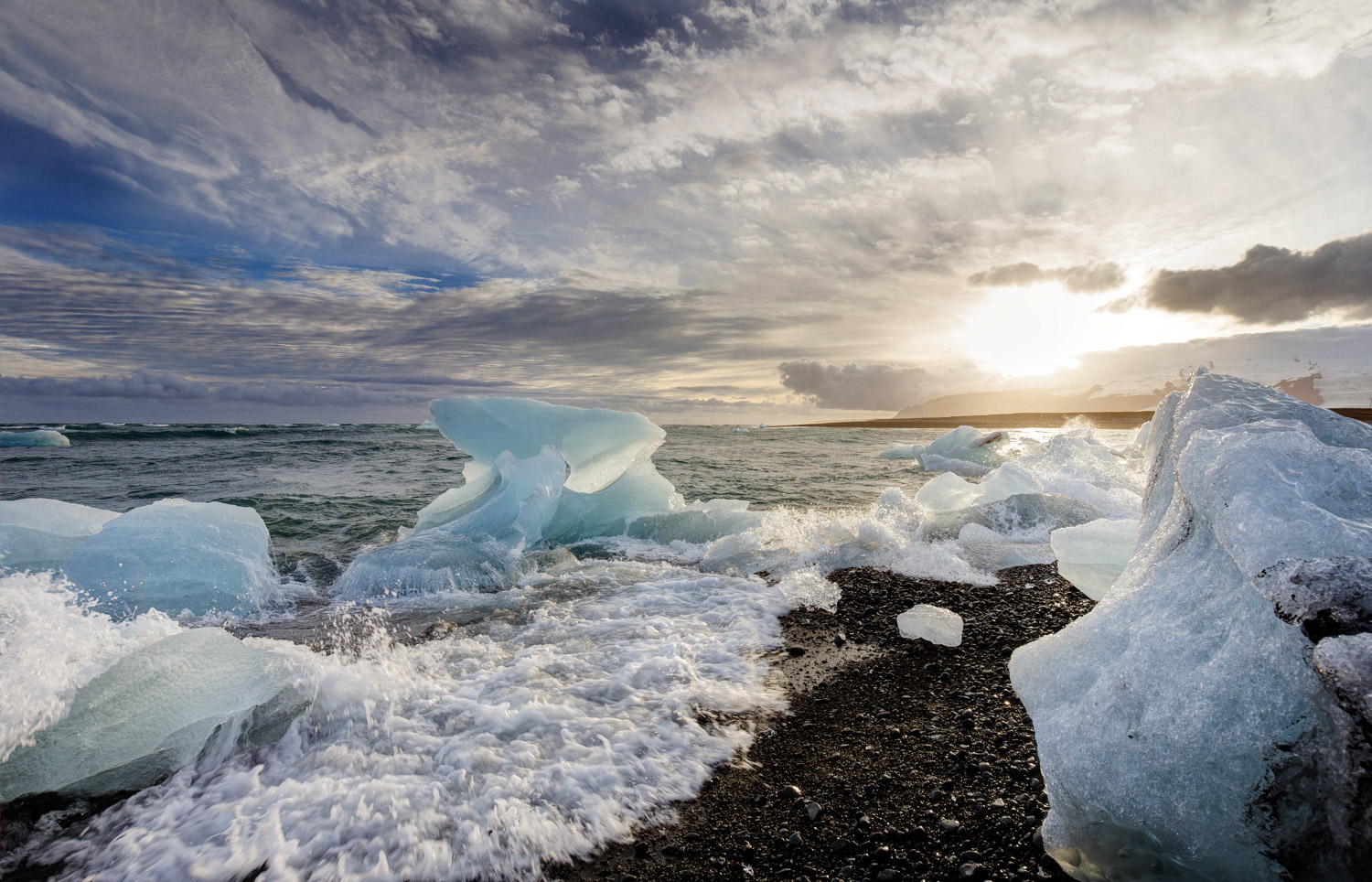 Eisbrocken am Diamond Beach in Island, Wellen brechen über schwarzen Kies, tief stehende Sonne und dramatischer Himmel im Hintergrund.