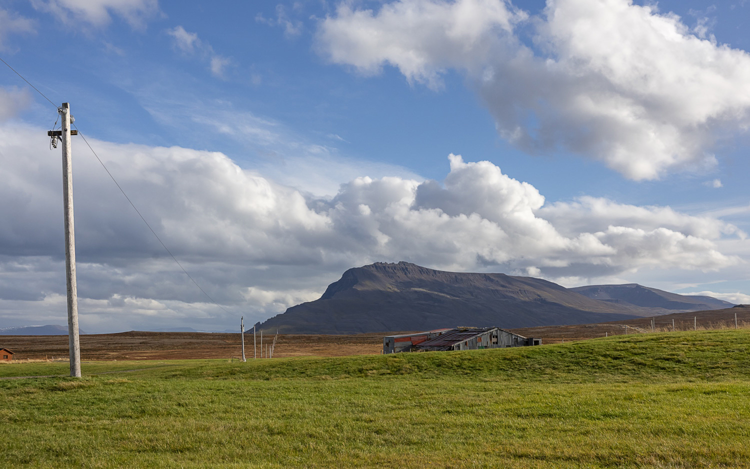 Weite Landschaft in Nordisland mit grünem Grasland, alter Scheune und Strommasten vor einem flachen Bergmassiv unter wolkigem Himmel.