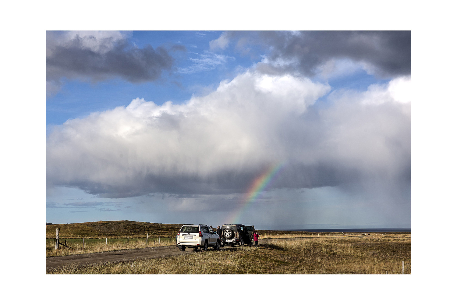 Fotostopp in Nordisland: zwei Geländewagen am Schotterweg, weite Landschaft und ein Regenbogen unter dramatischen Wolken über dem Meer.