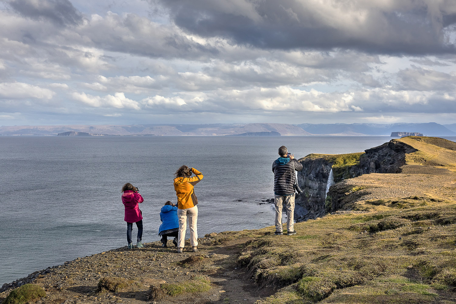 Fotografen an einer Klippe in Nordisland mit Blick über das Meer, dramatische Wolken und ein Wasserfall stürzt rechts die Steilküste hinab.