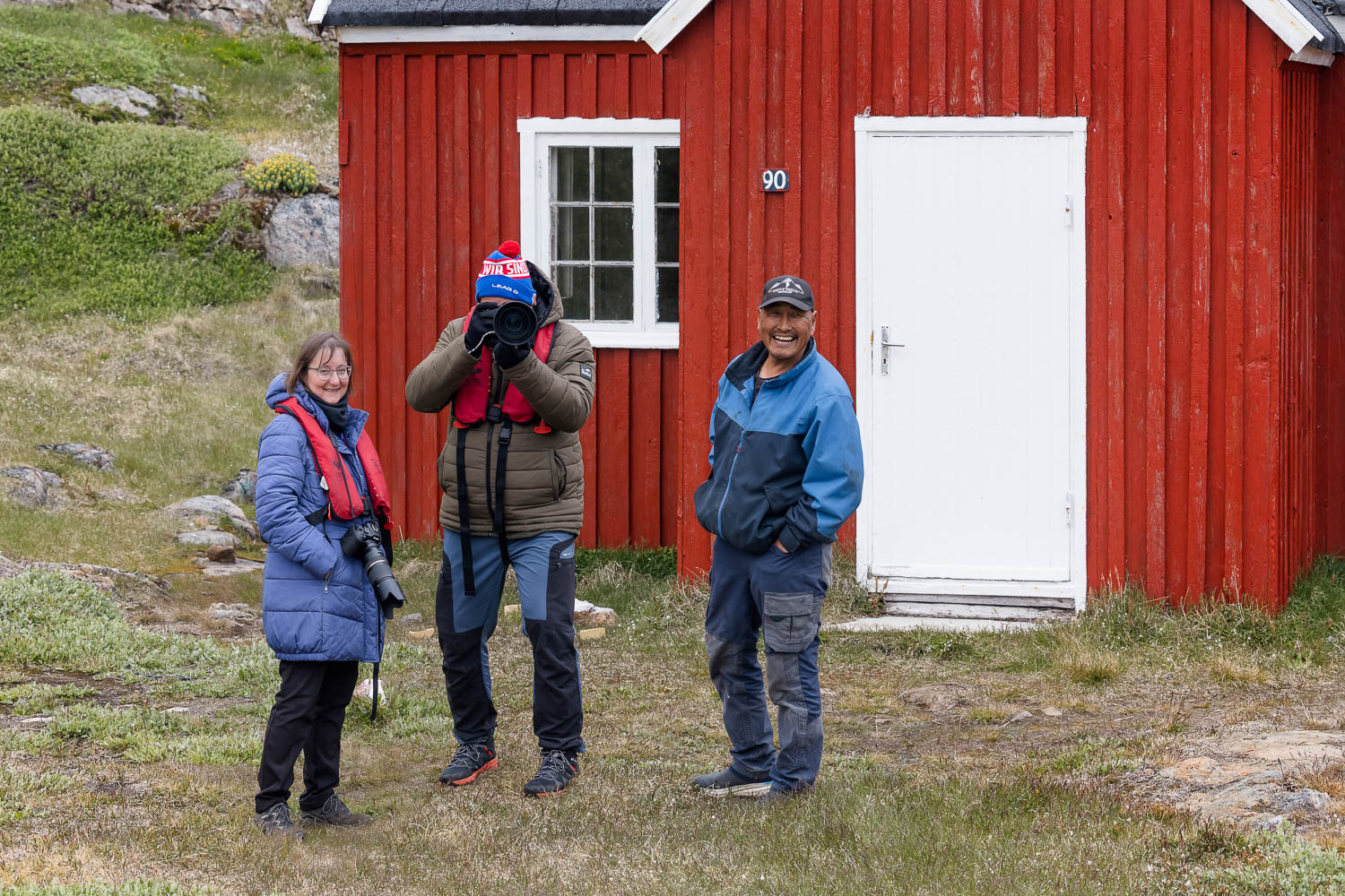 Fotoreise Ostgrönland mit Spaß Gut gelaunte Teilnehmer einer Fotoreise mit Kamera vor einem roten Haus in Ostgrönland Foto: Radmila Dier