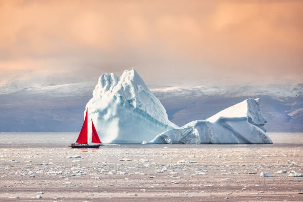 Segelboot mit roten Segeln vor einem gewaltigen Eisberg in der Diskobucht bei Ilulissat, Grönland.