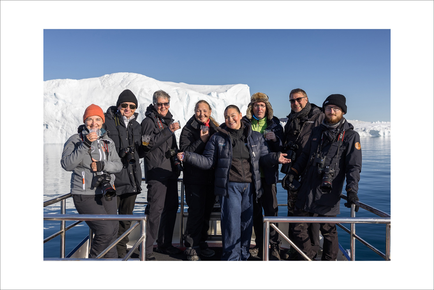 Fotoreise-Gruppe an Bord eines Bootes in der Diskobucht bei Ilulissat, Fotografen mit Kameras stoßen vor treibenden Eisbergen auf das gemeinsame Erlebnis in Grönland an.