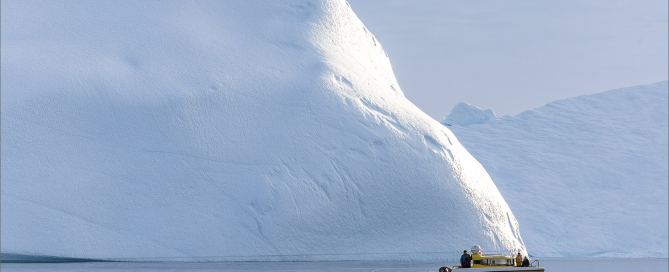 Kleines Ausflugsboot vor einem riesigen Eisberg in der Diskobucht bei Ilulissat, Westgrönland, ruhiges Wasser und arktische Weite.