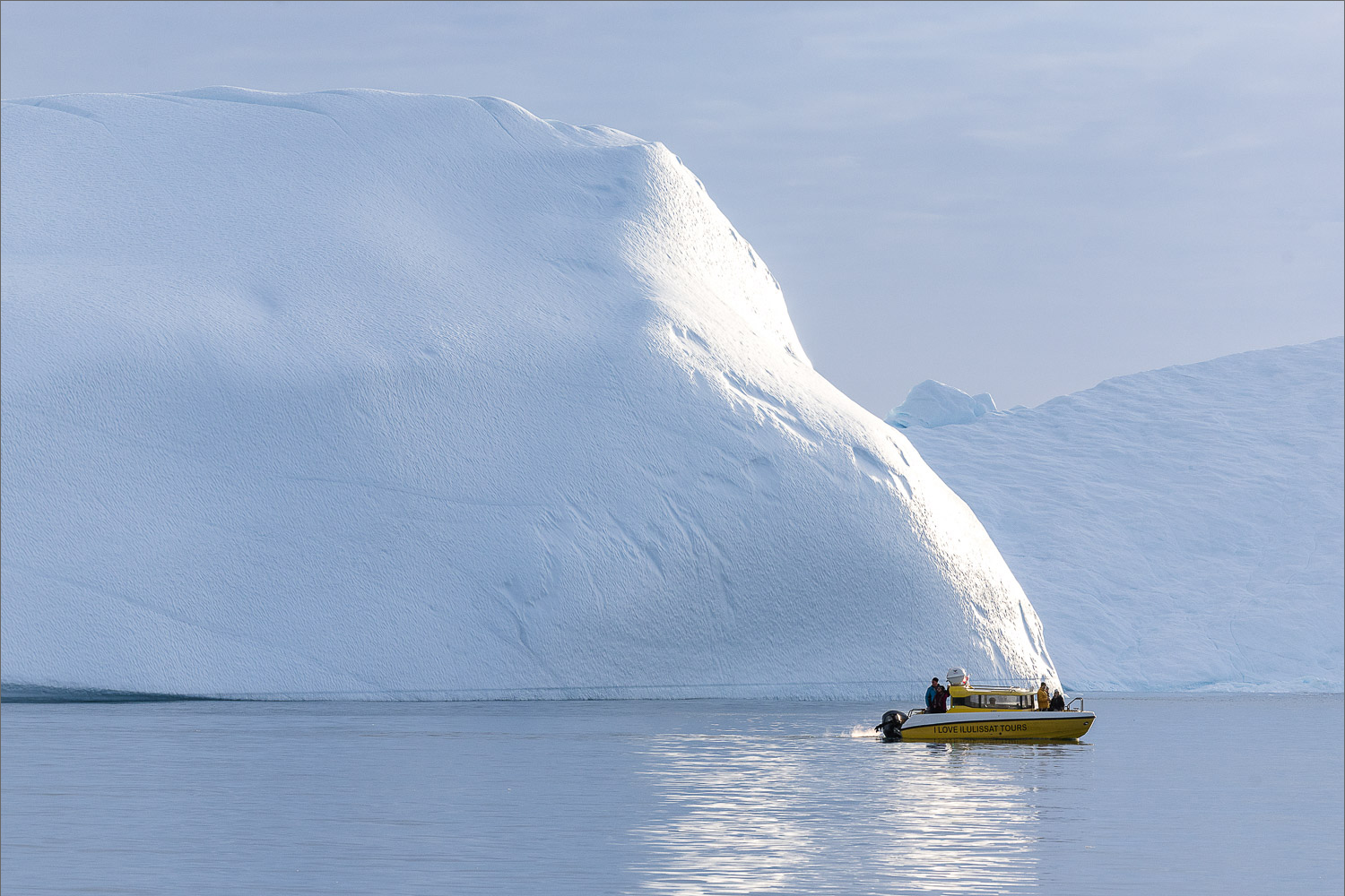 Kleines Ausflugsboot vor einem riesigen Eisberg in der Diskobucht bei Ilulissat, Westgrönland, ruhiges Wasser und arktische Weite.