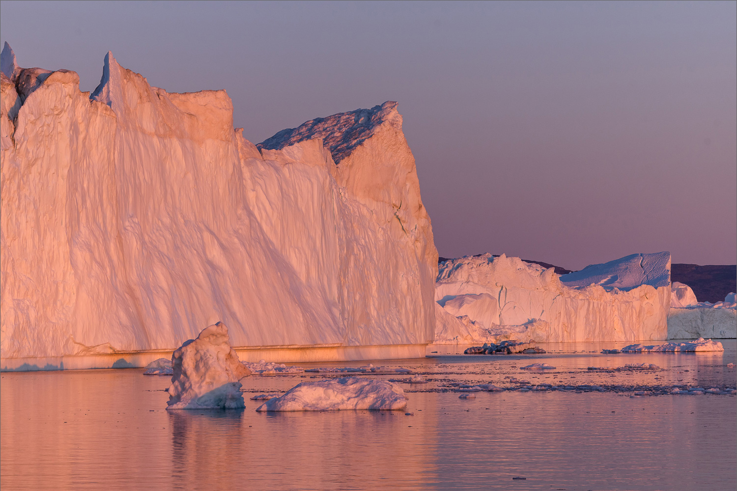 Große Eisberge in der Diskobucht bei Ilulissat im warmen Abendlicht, gespiegelt im ruhigen Wasser während der Blauen Stunde in Grönland.
