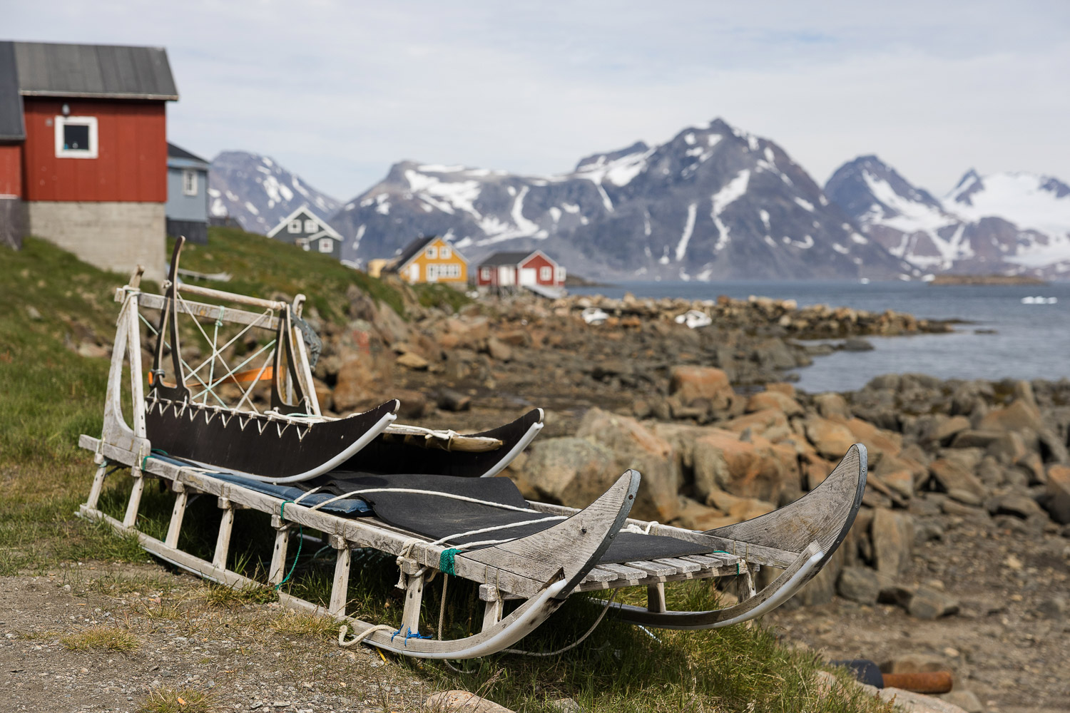 Hundeschlitten in Kulusuk Traditionelle Hundeschlitten an der Küste von Kulusuk mit bunten Häusern und Bergen im Hintergrund