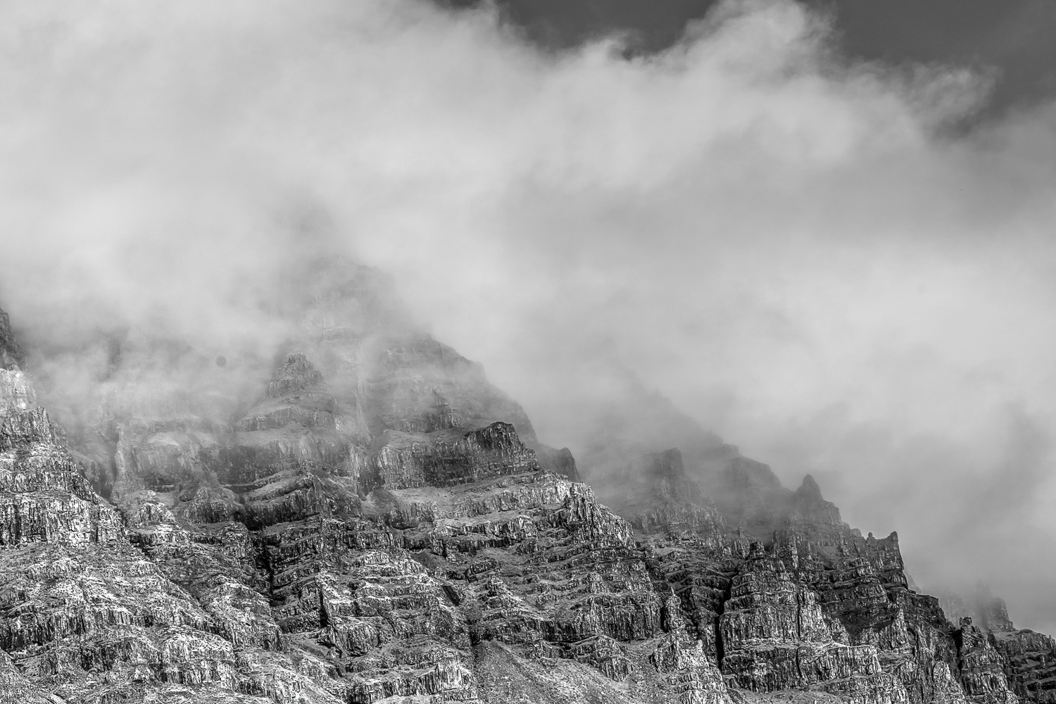 sland Landschaft (S/W) – Felswand im Nebel und tiefen Wolken Schwarzweißaufnahme einer zerklüfteten Felswand in Island, teilweise von tief hängenden Wolken und Nebel verhüllt.