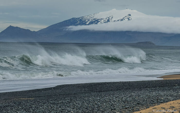 Brechende Wellen mit verwehter Gischt vor einem Kieselstrand auf Snæfellsnes; im Hintergrund der schneebedeckte Snæfellsjökull mit Wolkenband.