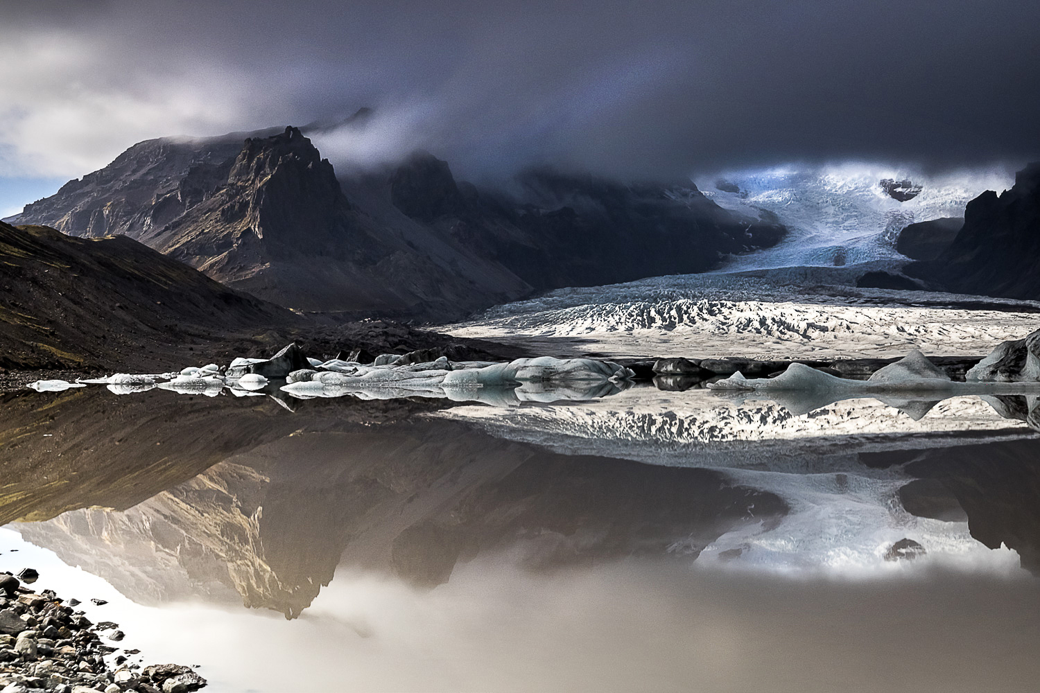 Island Gletscherlagune: Spiegelung von Gletscher und Bergen im Sturmlicht Gletscherlagune in Island mit Eisbergen und starker Spiegelung von Bergen und Gletscher, dramatische dunkle Wolken hängen tief über der Landschaft.