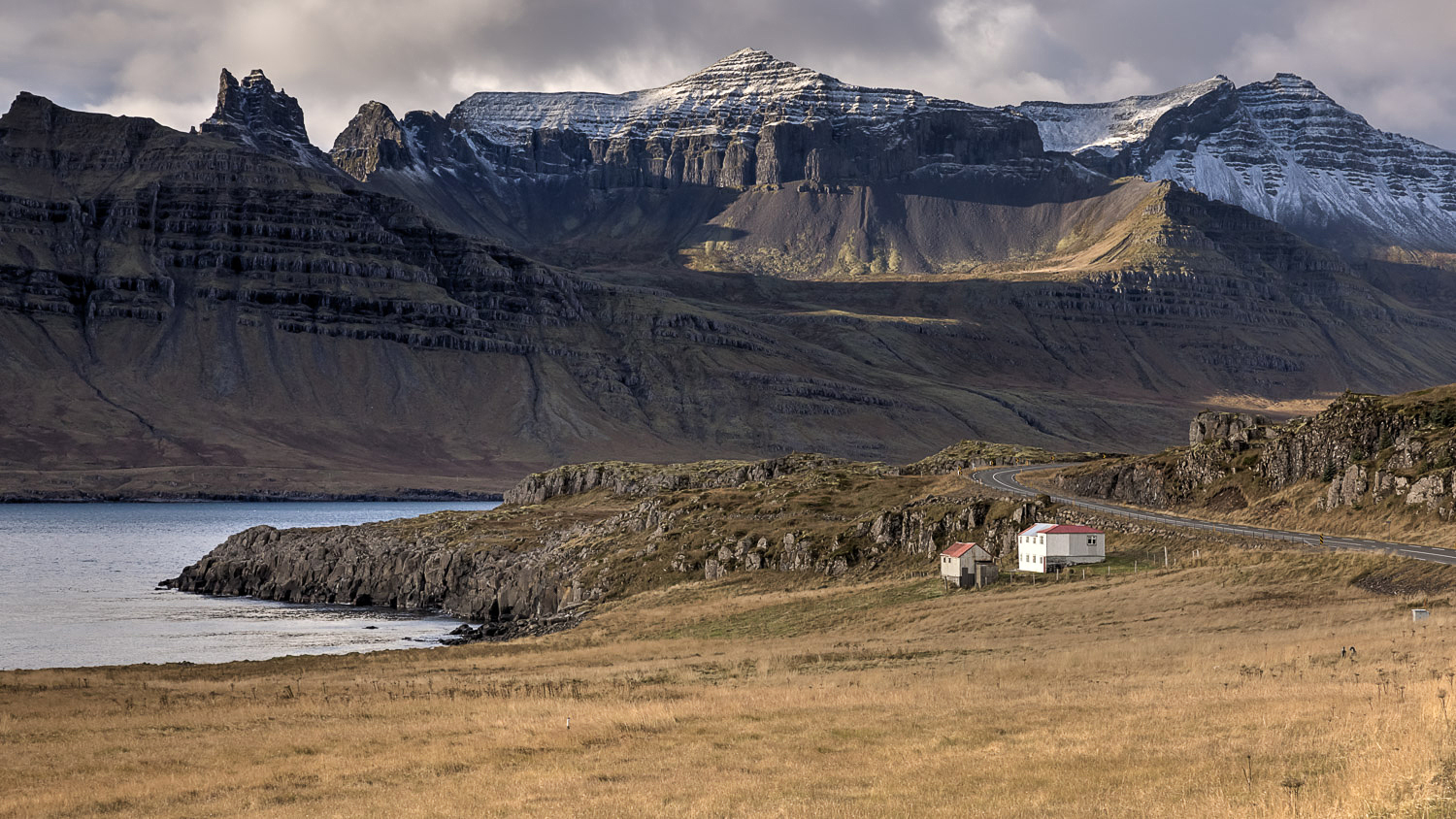 island-ostfjorde-fjord-kueste-berge-strasse-herbstlicht Fjordküste in Islands Ostfjorden mit kurviger Straße und kleinen Häusern, dahinter mächtige Berge mit ersten Schneefeldern unter dramatischem Himmel.