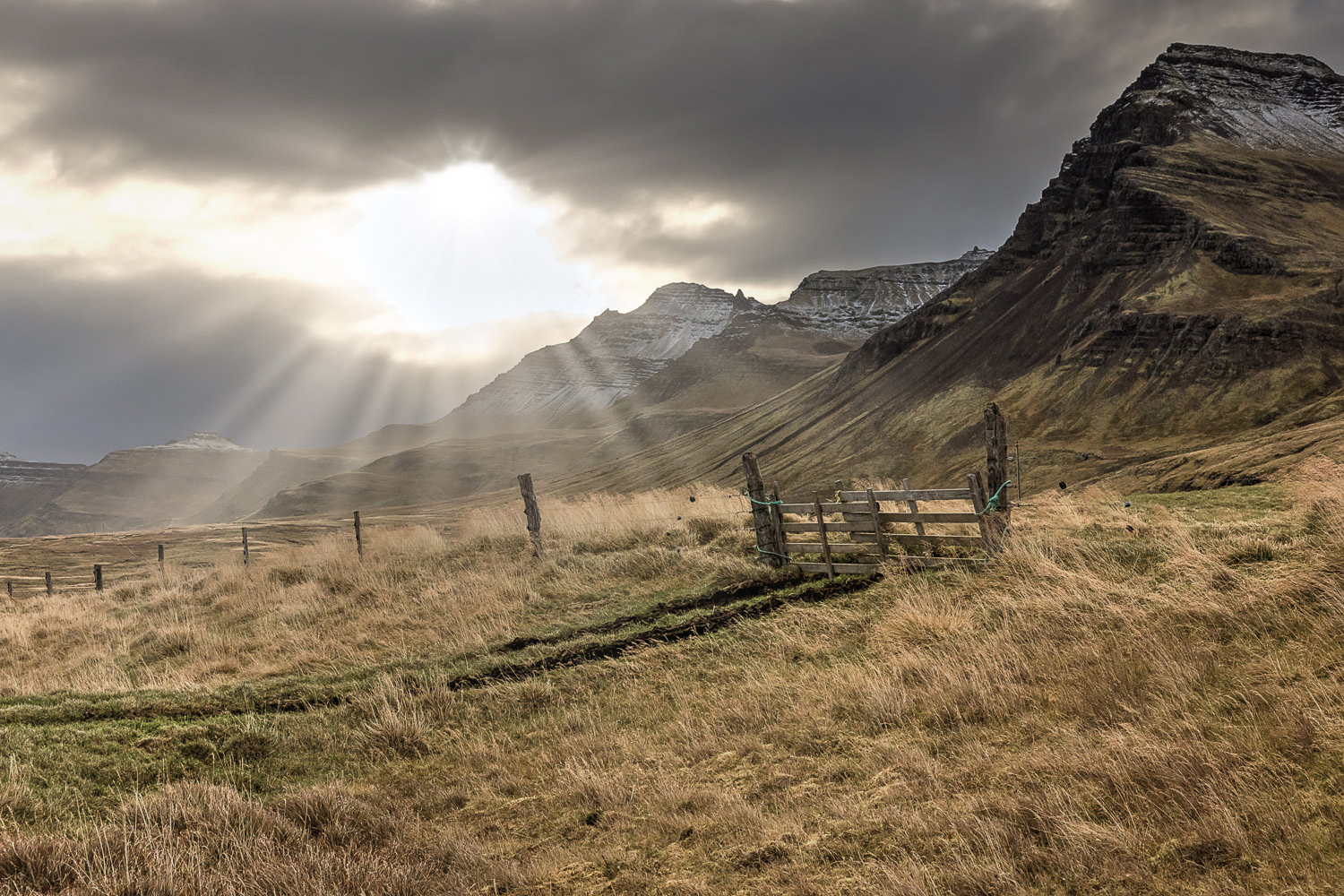 island-ostfjorde-lichtstrahlen-berge-herbstlandschaft Sonnenstrahlen brechen durch Wolken über steilen Bergen in den Ostfjorden in Island, Vordergrund mit herbstlichem Gras und Zaun.