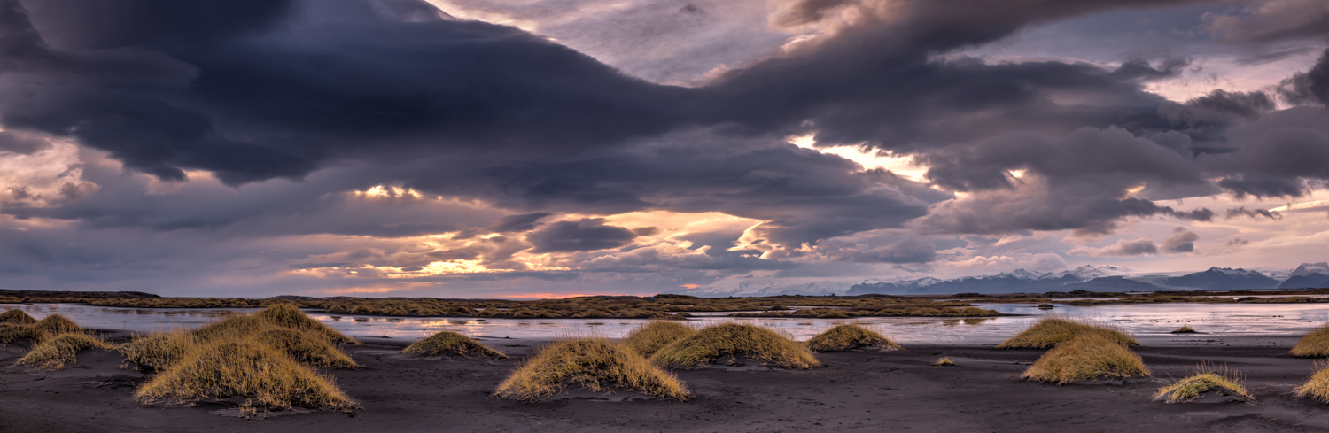 Panorama mit schwarzem Sand und grasbewachsenen Dünen bei Stokksnes in Island, dramatischer Himmel im Abendlicht und Berge am Horizont.