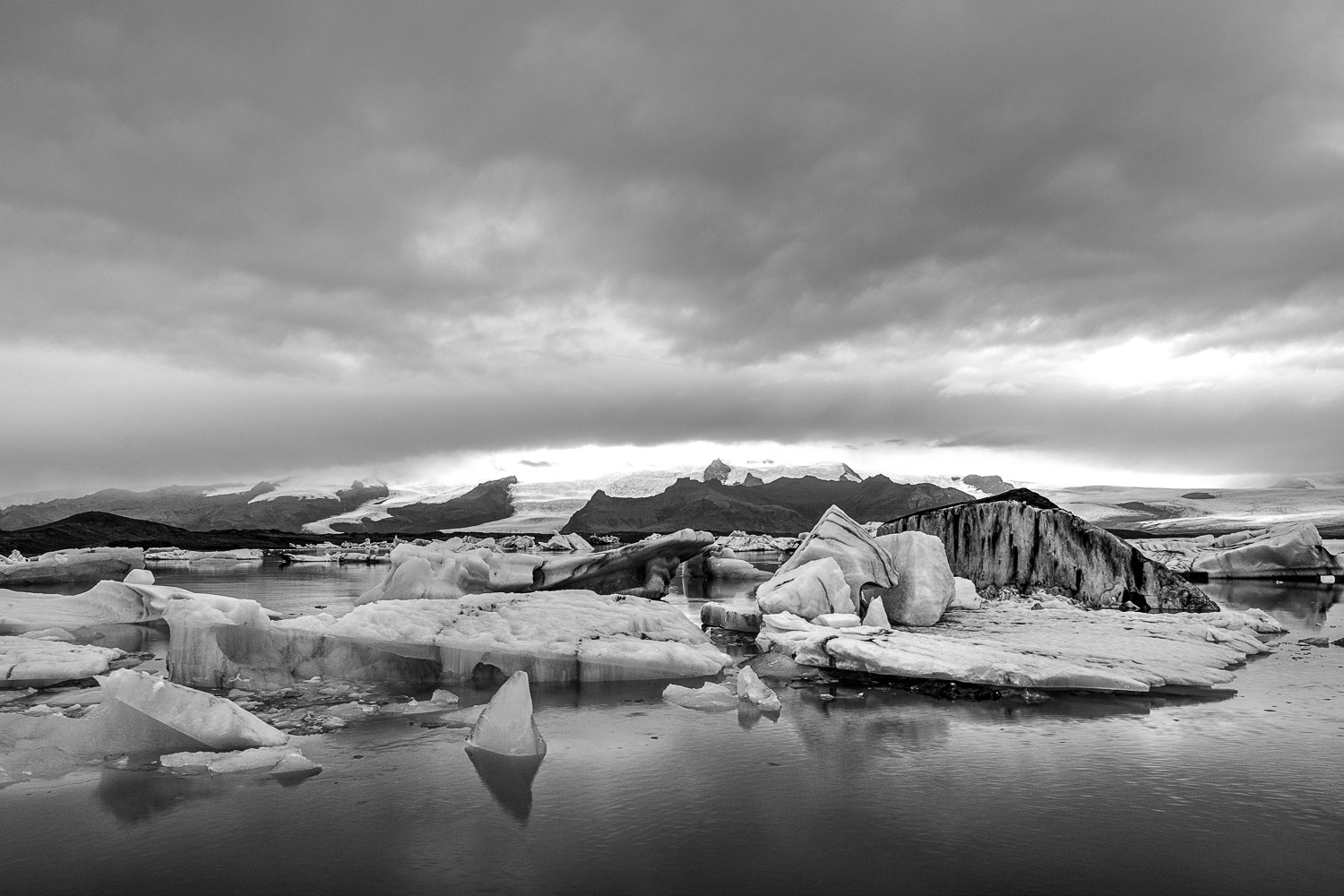 Jökulsárlón Island (S/W) – Eisberge in der Gletscherlagune Schwarzweißfoto der Gletscherlagune Jökulsárlón in Island: Eisberge im ruhigen Wasser, dahinter Gletscher und dramatische Wolken.