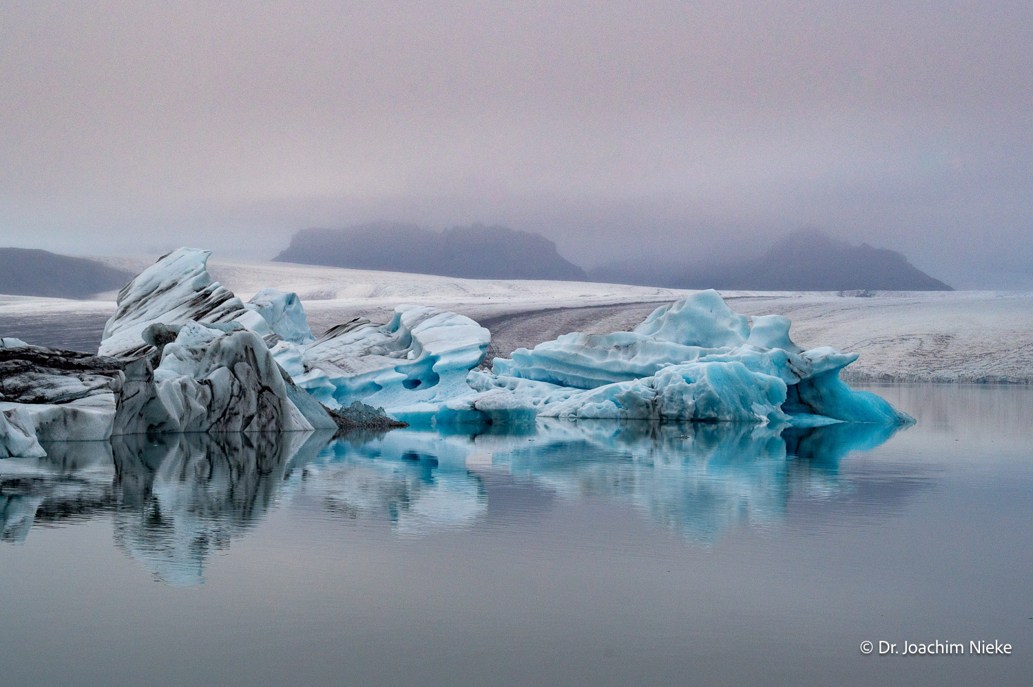 Blaue Eisberge spiegeln sich im ruhigen Wasser der Gletscherlagune Jökulsárlón unter tiefen Wolken
