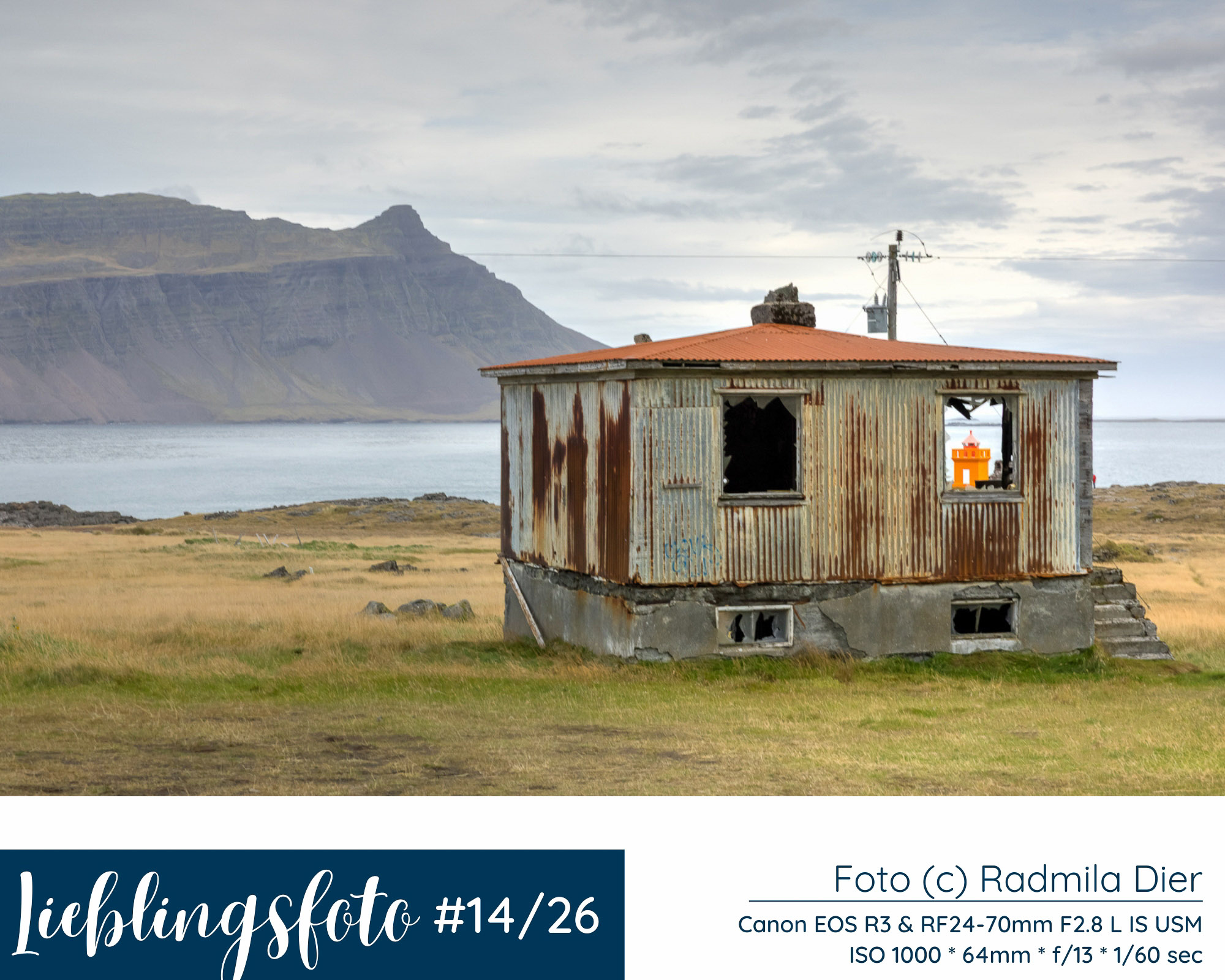Verlassenes Wellblechhaus am Fjord in Island mit Bergen im Hintergrund und orangefarbenem Leuchtturm im Fenster