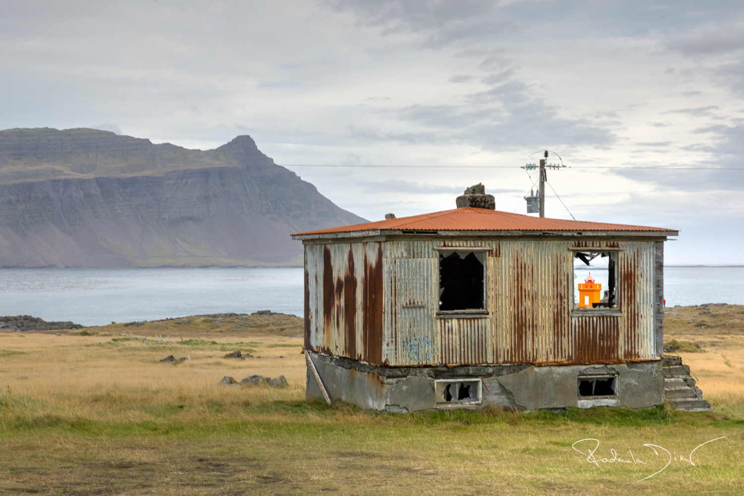 Verlassenes Wellblechhaus in Island mit Blick aufs Meer und Berge, im Fenster ist ein orangefarbener Leuchtturm zu sehen.